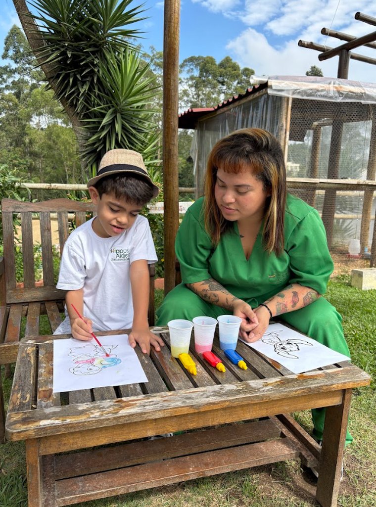 Criança pintando ao lado de uma profissional da Hippus Aldeia em mesa ao ar livre